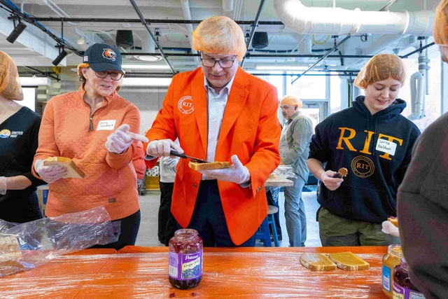 RIT President Bill Sanders and his wife, Emily, spread jelly on a piece of bread. A student stands next to them spreading peanut butter on the pre-jellied bread. All of them wear RIT orange hairnets.