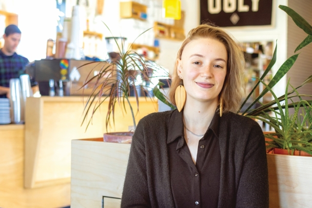 a college age student with mid-length blond hair and a black shirt sits in front of a plant in an office environment.