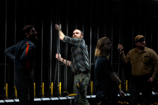A man shows how to lower and raise set pieces behind the scenes of a theater as students look on.