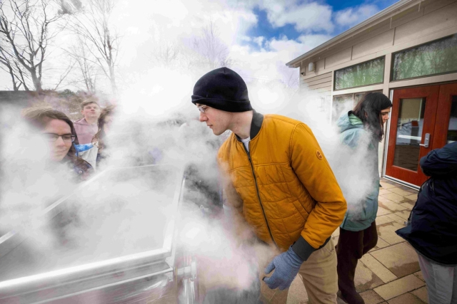 Man in yellow jacket leans into steaming container as students gather outside building.