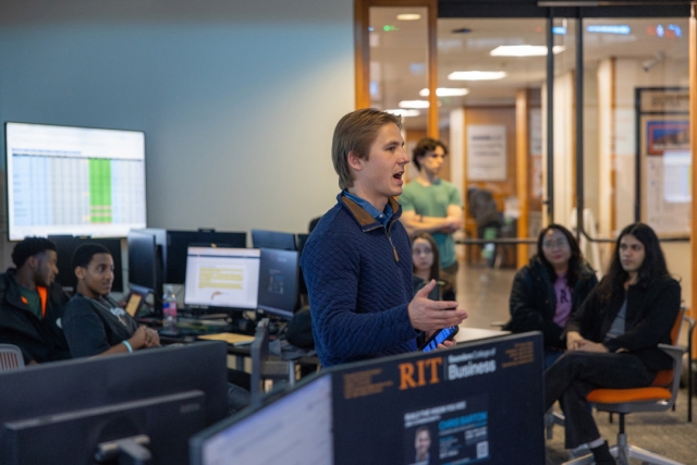 a young man stands in a finance lab speaking to a club