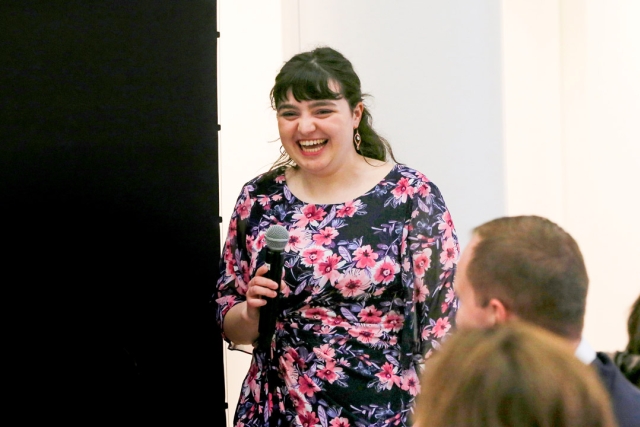 a studeny in a floral dress and long brown hair speaks in to a microphone at an event