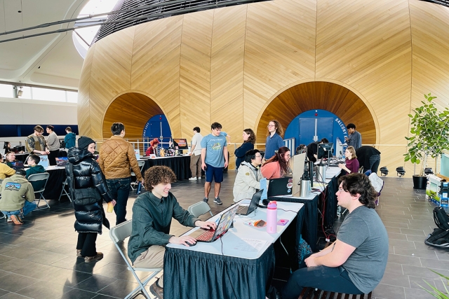 Students sit across from each other at tables in a college atrium where laptops are setup for game playtesting