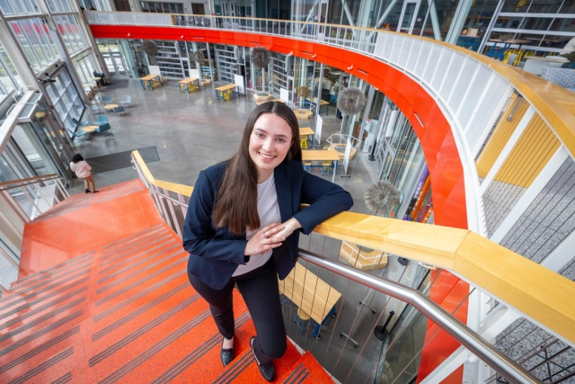 a student stands in an orange and white staircase in a large, bright, modern space.