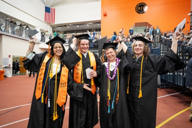 Four college graduates in an arena smile and cheer while wearing black caps and gowns.