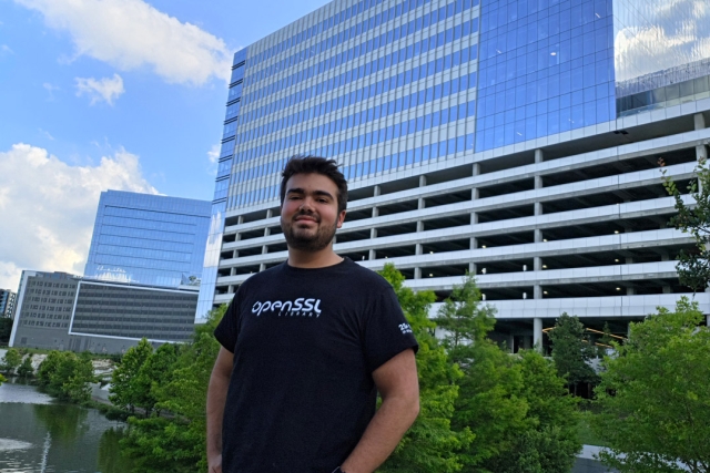 A young man in a black tshirt stands in a park like area with city buildings behind him.