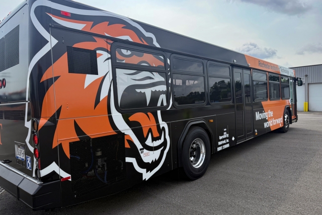 An image of the new RIT Bus. Looking at the side with the rear closest - it is black with an orange vertical stripe near the front and a tiger logo near the rear.