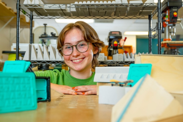 a collage age person with short hair and glasses looks at the camera through a shelving unit