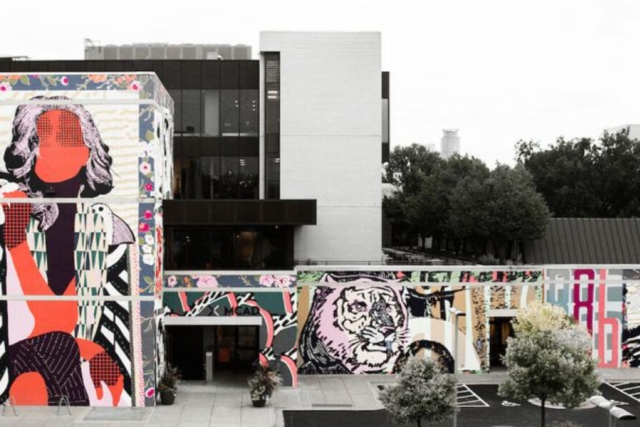 A photo of the exterior of an unidentified building, likely a college campus. The building is covered in mural art featuring a pop-art style female figure on the left, and a pop-art style tiger on the right. There are various abstract designs in cool tones and pinks in the background of the mural.