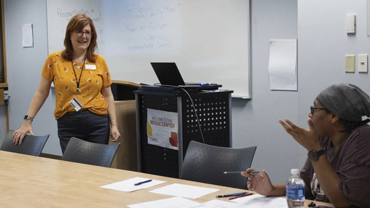 One student seated at a table, talking with a faculty member.