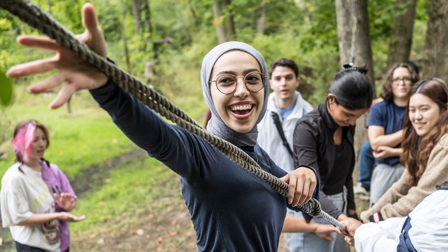 A student reaches out to grab a rope while other students hold onto the rope in the background during an outdoor teambuilding event.