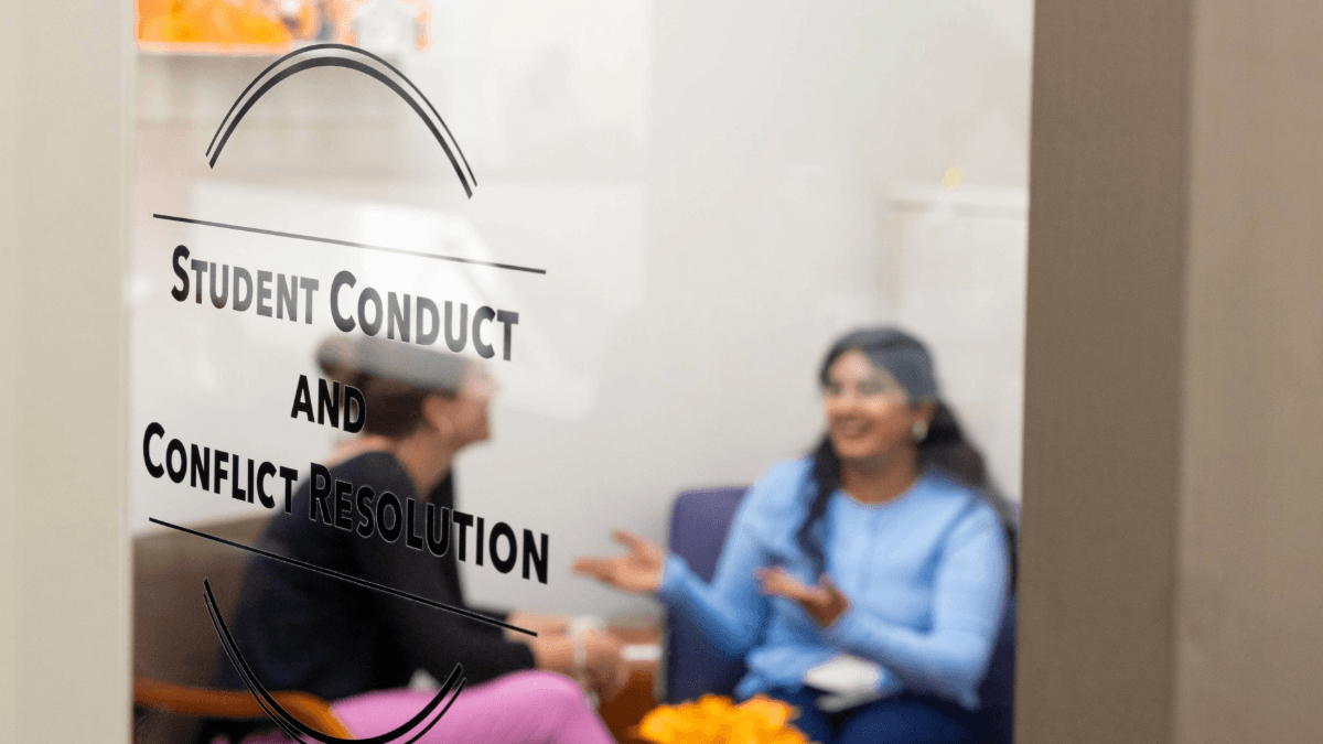 A student and staff member sitting inside an office - a clear door in the foreground labeled "Student Conduct and Conflict Resoultion"