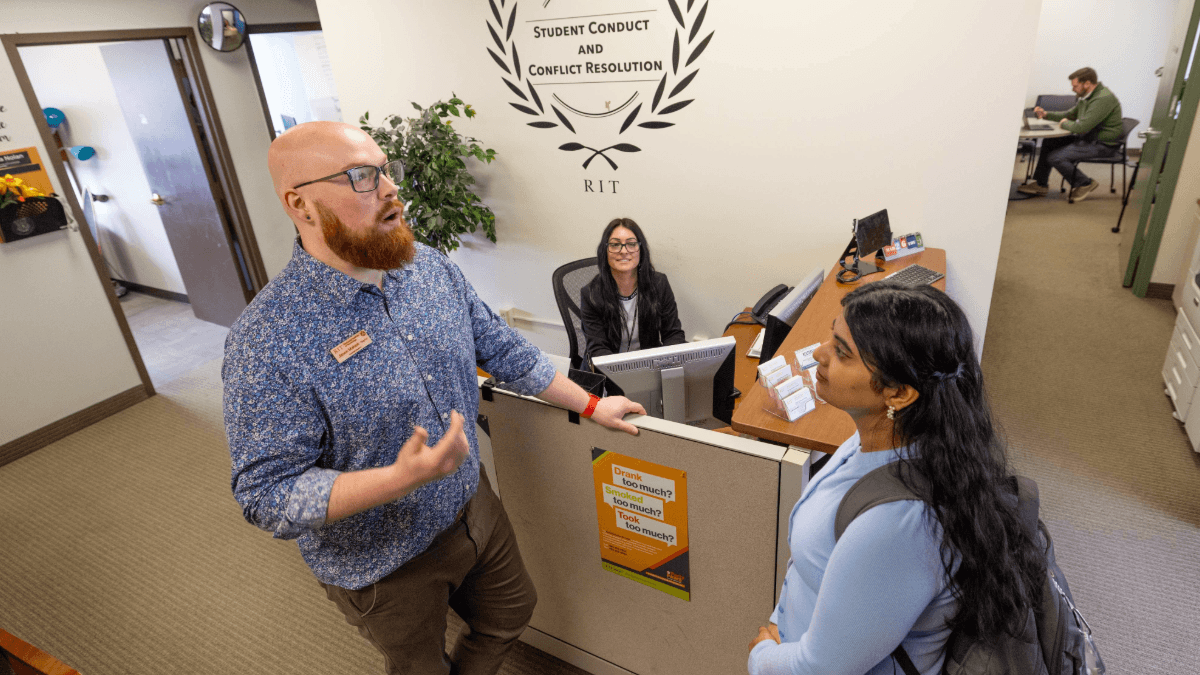 A student speaks with a staff member at the RIT Student Conduct and Conflict Resolution office while another staff member works at a desk in the background