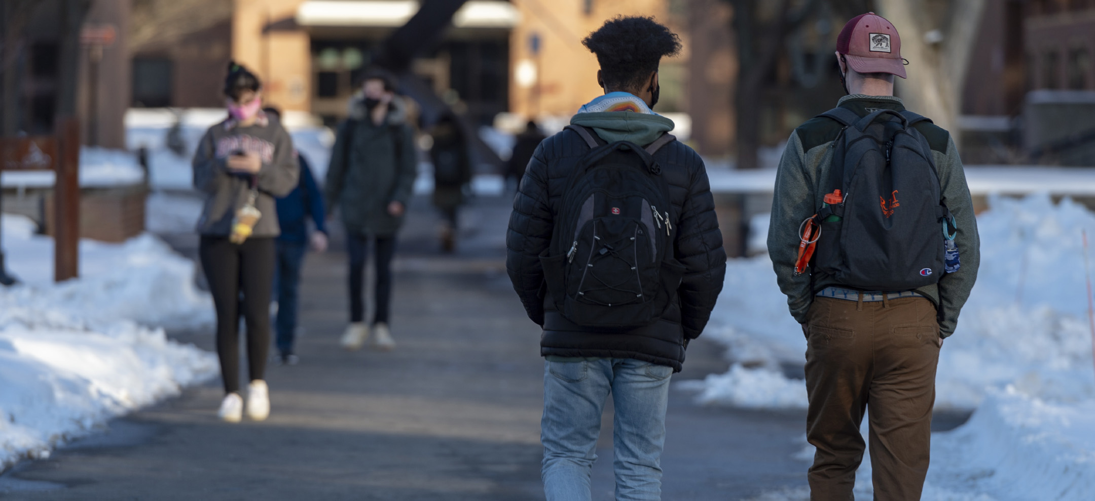 An image of two RIT students, walking down the Quarter Mile, towards the Sundialand the dorms. It is wintertime and the sidewalks are shoveled and there are snow mounds beside the pathway.