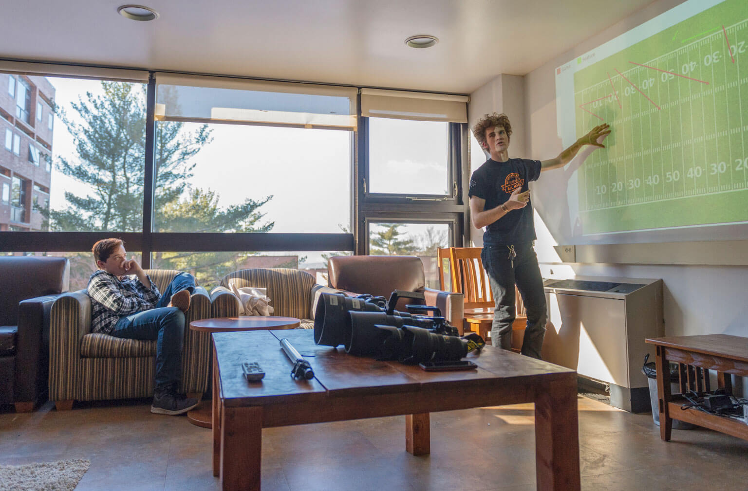 An image of two RIT students in a dorm lounge, one sitting and the other standingand pointing at a projection of a football field on a screen. Behind the students is a wall offloor-to-ceiling windows and in front of the students is a coffee table with large varying sizes of camera lenses.
