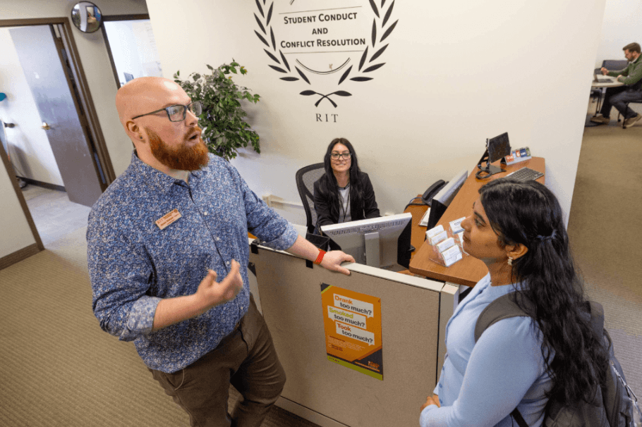 A student speaks with a staff member at the RIT Student Conduct and Conflict Resolution office while another staff member works at a desk in the background.