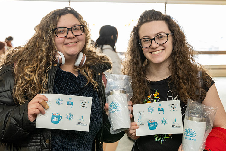 Two students pose smiling while holding FreezeFest stickers and FreezeFest tumbler mugs
