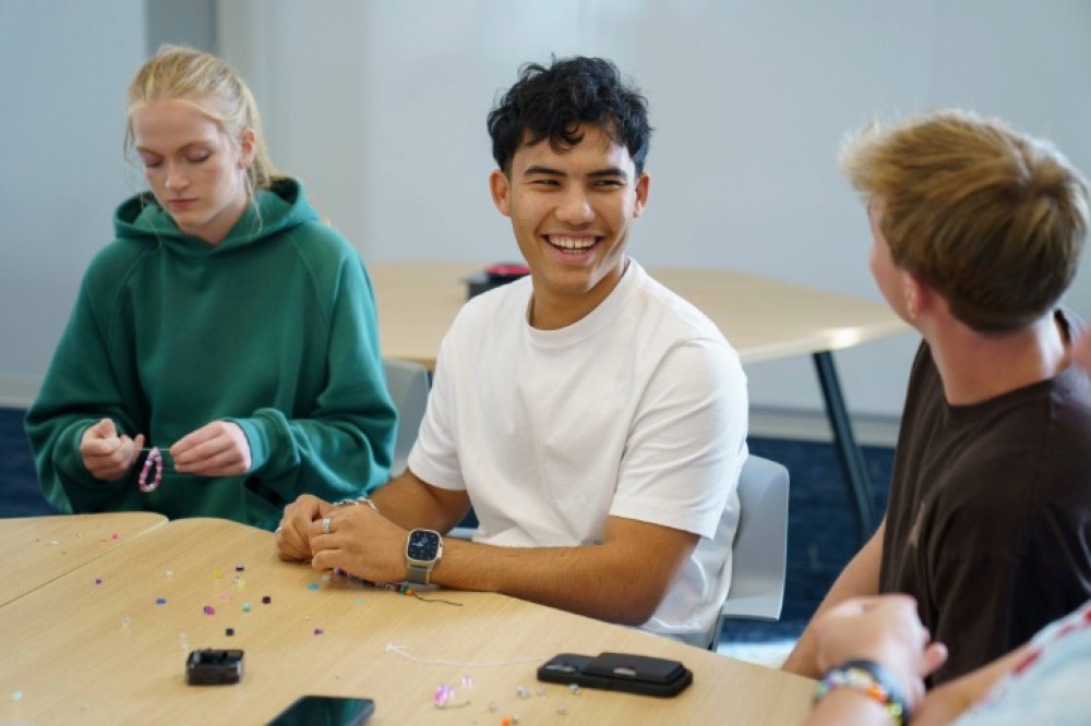 Three students sit around a table indoors, stringing colorful beads into bracelets, with small beads and craft supplies scattered across the table.