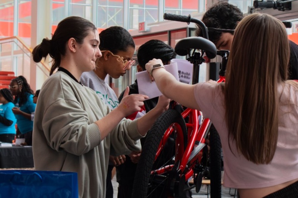 Students work together at a table assembling a red bicycle, using tools and following instructions during a group activity.