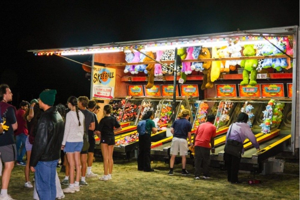 Students standing around a brightly lit skee-ball carnival booth at night, playing games and chatting beneath colorful lights and rows of hanging stuffed prizes.