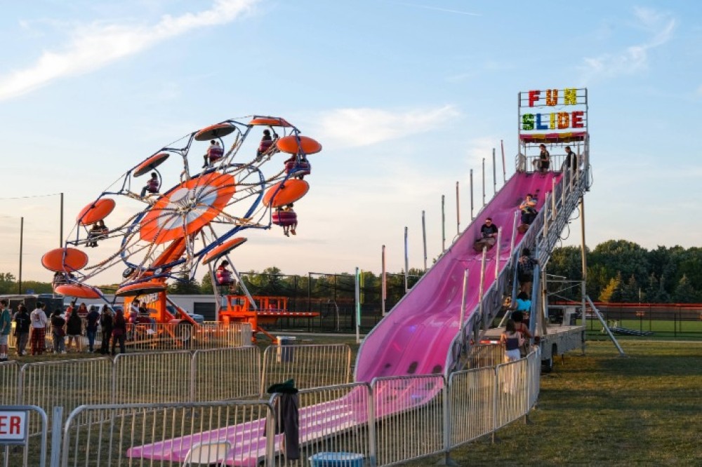 Students gather around outside a carnival ride and a tall pink “Fun Slide” on a grassy field under a blue sky.