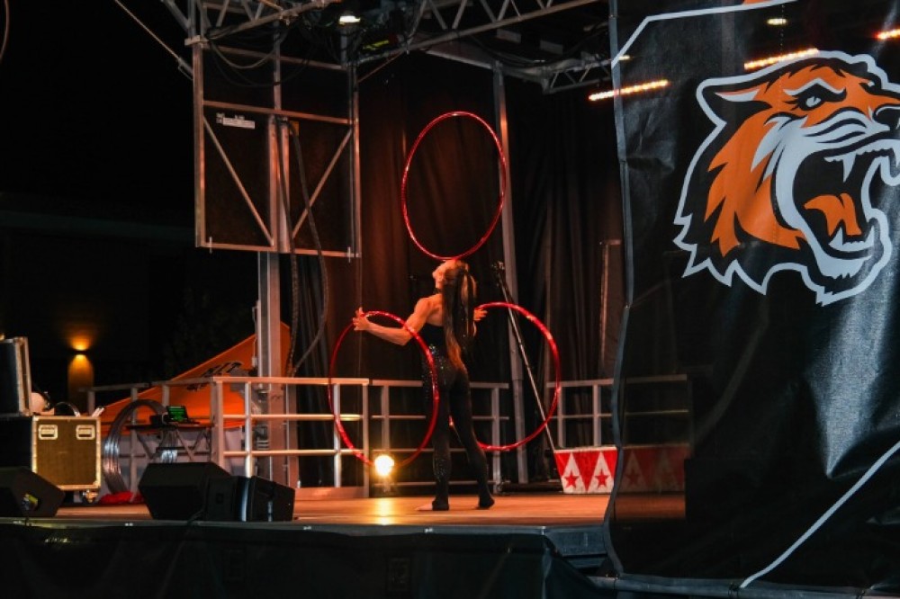 A performer stands on an outdoor stage at night, spinning balancing rings, with stage equipment and a large RIT tiger logo visible nearby.