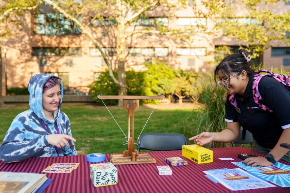 Two students stand across from each other at an outdoor table playing a washer toss game, with small game boxes, flyers, and campus greenery visible in the background.