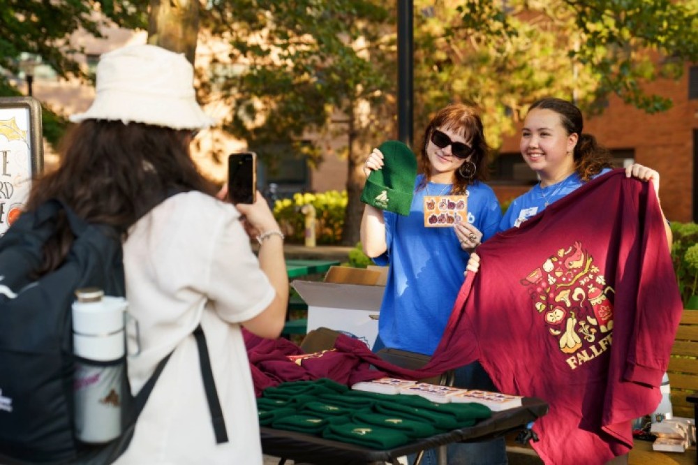 Students stand behind a table outside displaying apparel and accessories, holding up a green hat and a maroon T-shirt while another student takes a photo.
