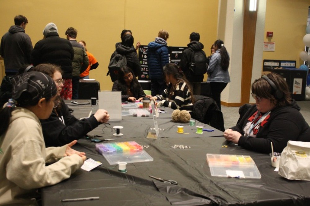Students sit around a large indoor table covered with supplies, threading beads and assembling crafts, while other students browse activity tables in the background.