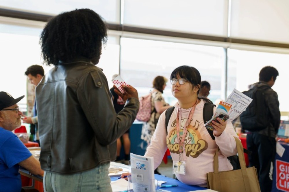 Students talk with a representative at an indoor campus event table, with other tables and attendees in the background.