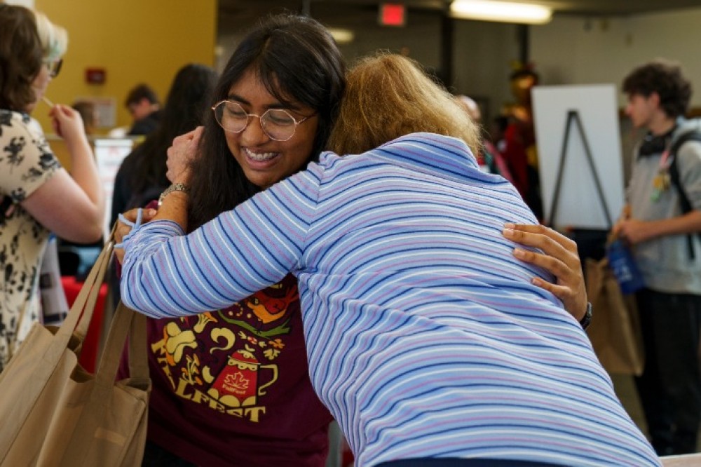 Two students hug inside a busy campus event space, surrounded by tables, flyers, and other attendees.