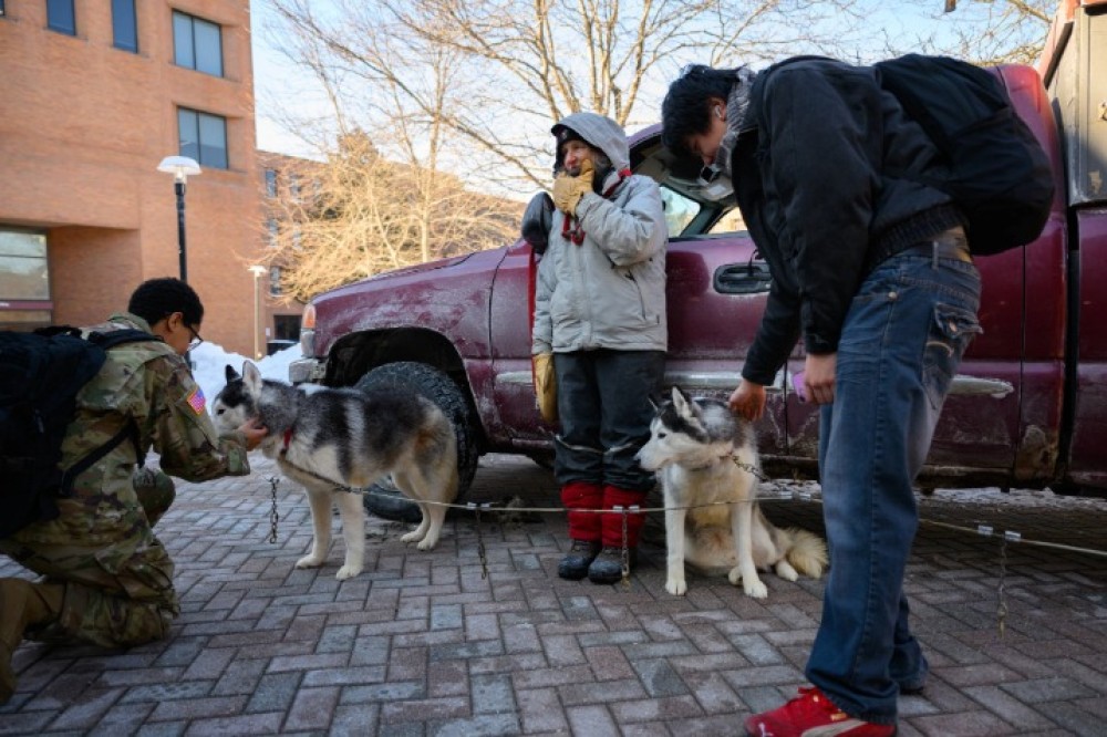 Students stand outside on a snowy campus walkway near a parked truck, interacting with two harnessed husky dogs while a handler stands nearby wearing winter gear.