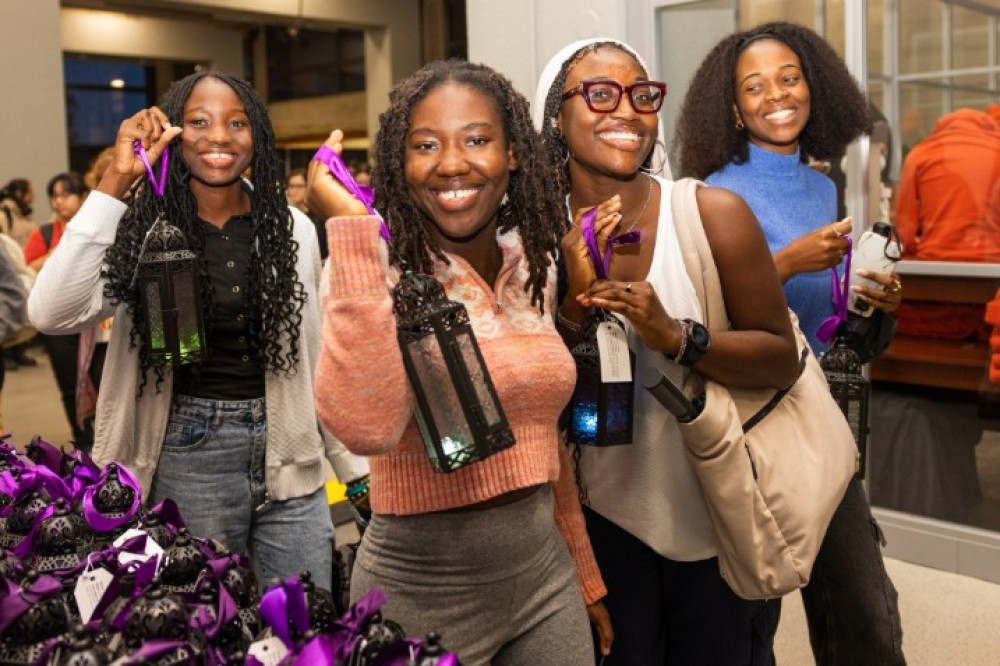 Students hold illuminated decorative lanterns with purple ribbons inside a campus space, standing near a table filled with identical lanterns.