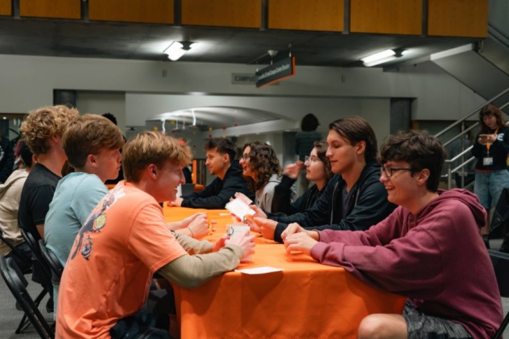 Students sitting and talking across from each other at a long table covered with an orange tablecloth indoors.