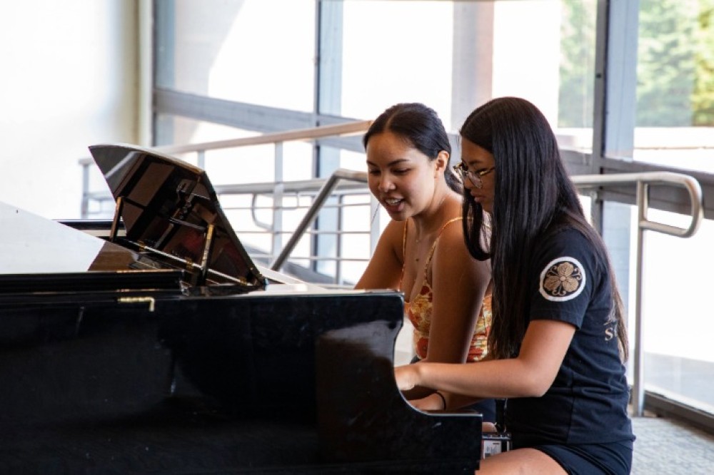 Two students sit at a grand piano inside a campus building, playing together near large windows and a railing.