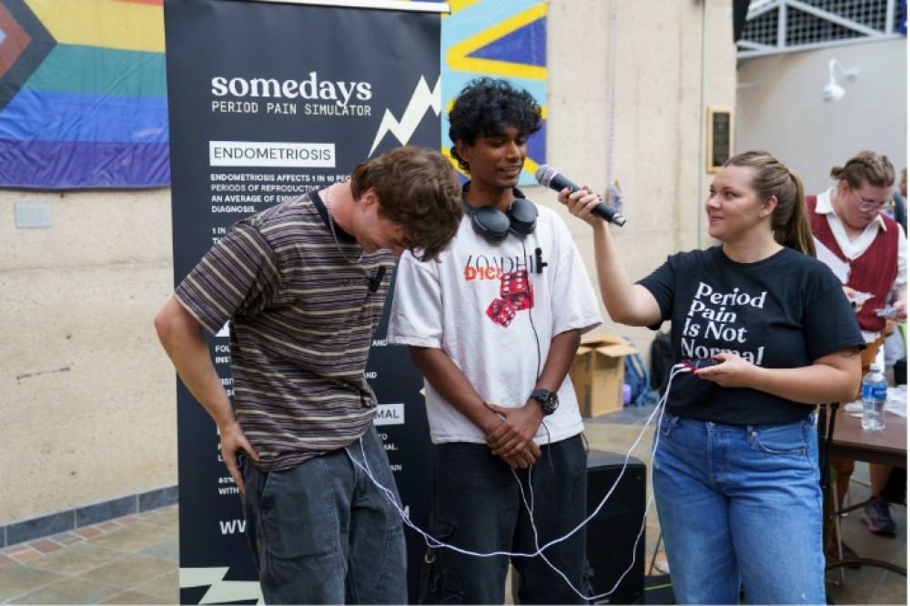 Students participate in an interactive indoor activity using a device connected by wires, while another person holds a microphone; a display behind them reads “Somedays Period Pain Simulator – Endometriosis” with informational text.