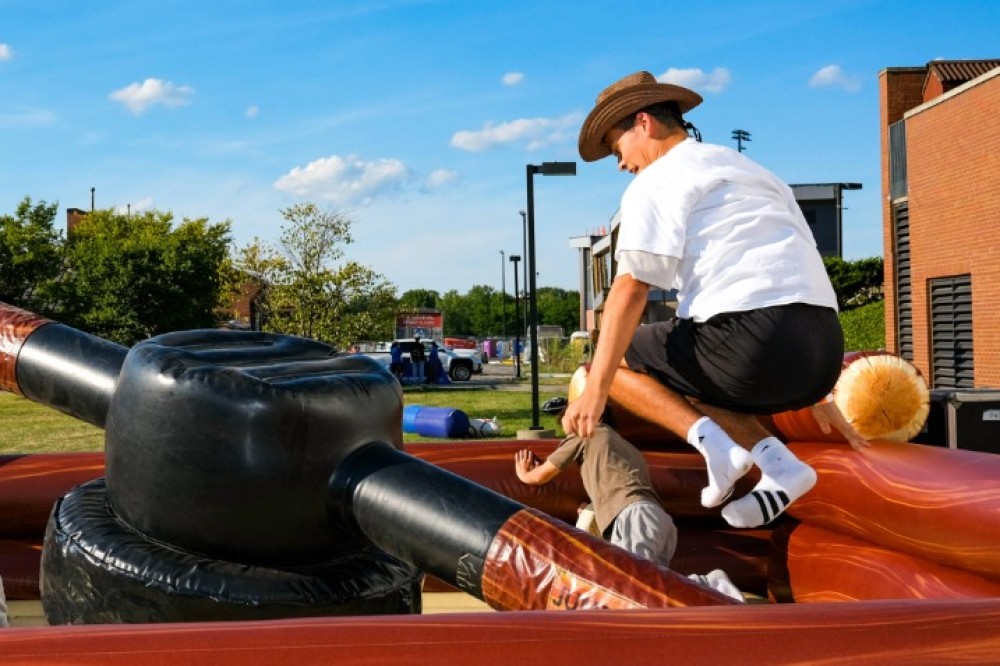 A student rides an inflatable mechanical bull outdoors on campus as others gather nearby, with buildings and open sky in the background.
