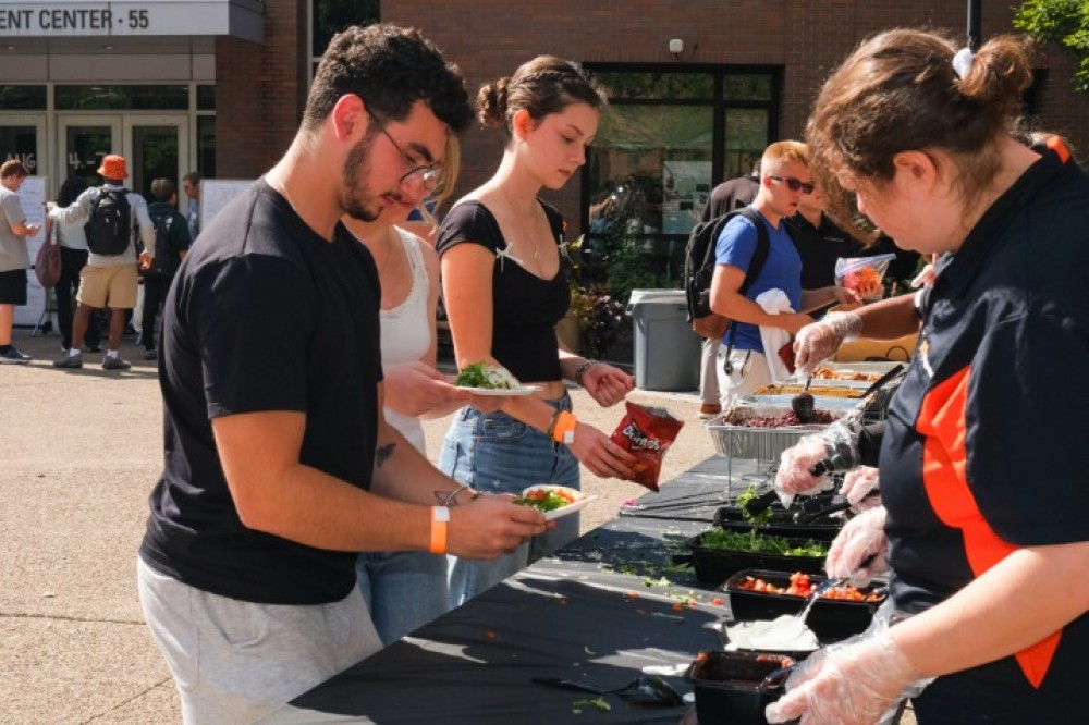 Students are served tacos at an outdoor station, adding toppings from trays while event staff assists behind a table.