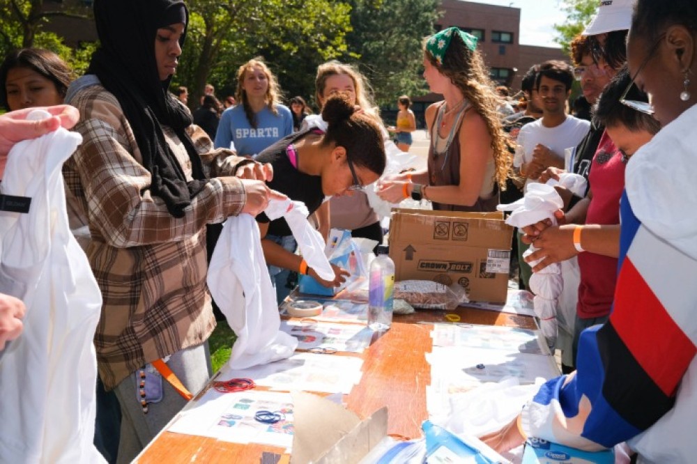 Students gather around an outdoor table filled with supplies, twisting and tying white T-shirts with rubber bands to prepare for tie-dye.