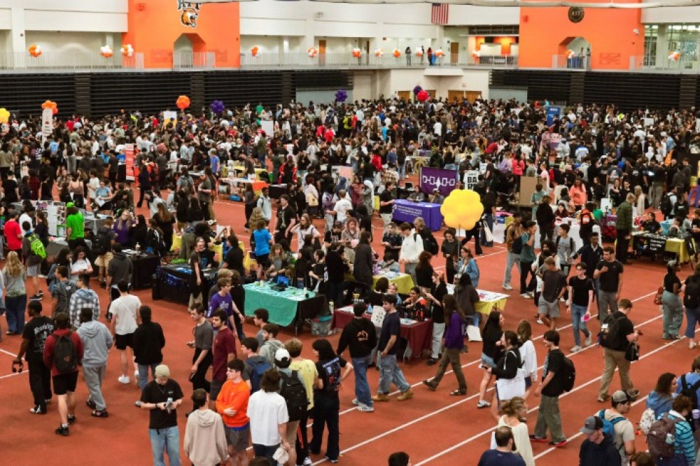 A large crowd of students moves through the indoor field house filled with tables and booths representing student organizations.
