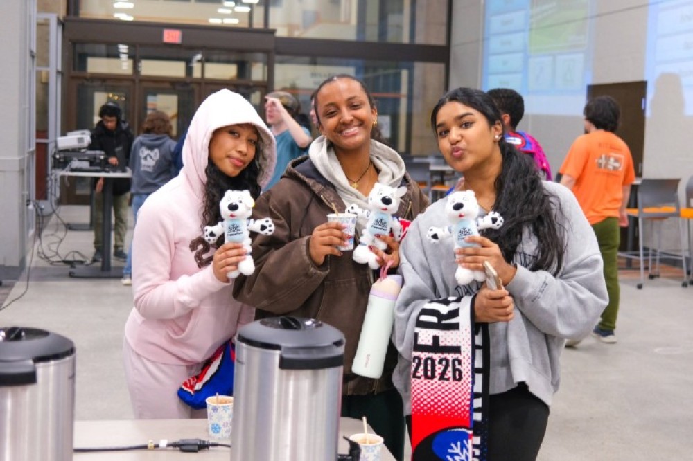 Three students stand indoors at a campus event, holding small stuffed Roarie plush toys and cups of hot beverages, with tables and other attendees visible in the background.