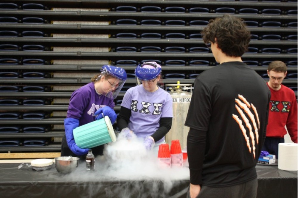 Students wearing face shields and gloves prepare liquid-nitrogen ice cream at a table, pouring ingredients into a container that releases visible white vapor, while others watch.