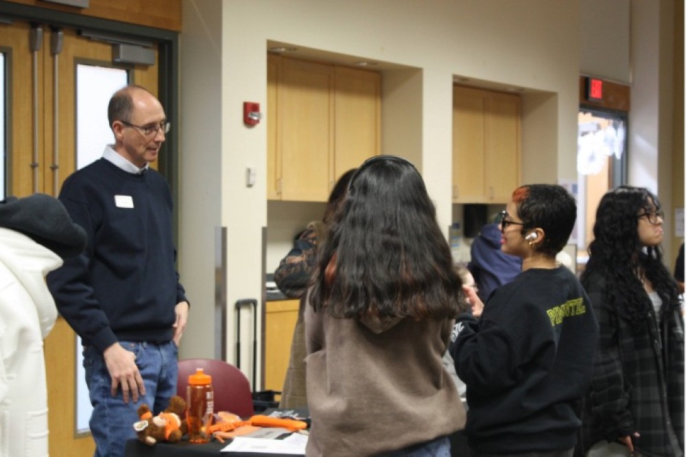 Students stand around an indoor table display speaking with a campus representative, with items such as water bottles, giveaways, and other informational materials.