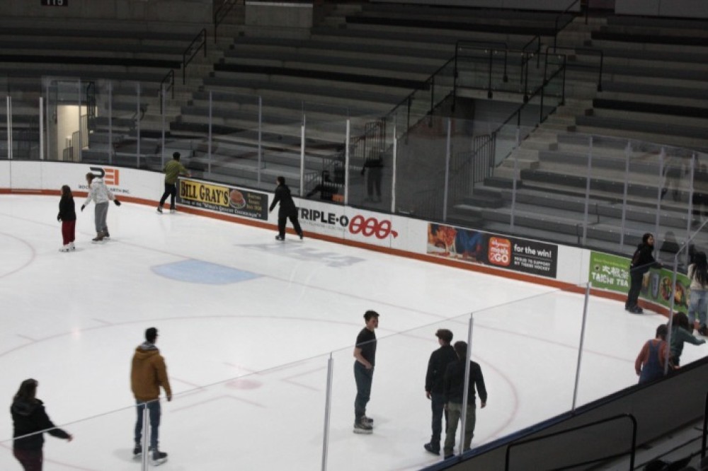 Students skate across an indoor ice rink, gliding along the boards while others stand and watch from nearby seating areas.