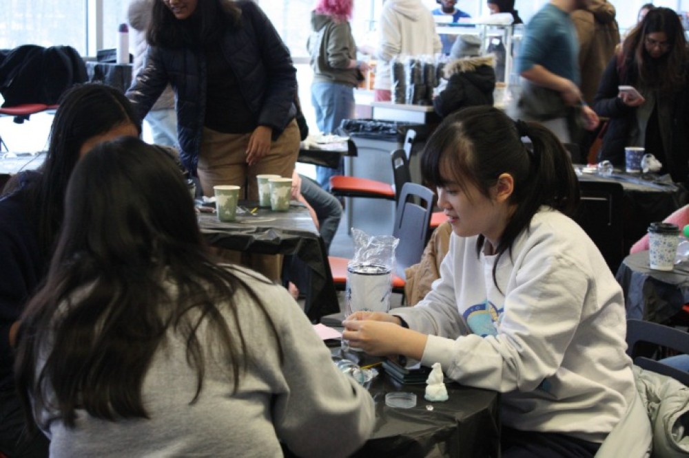 Students sit around an indoor table, working on a hands-on craft activity with small materials and tools, while others move through the event space in the background.