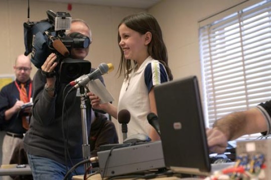 A young person speaks at a microphone while being filmed.