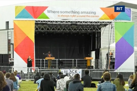 outdoor stage with person speaking at podium and students sitting in a lawn.