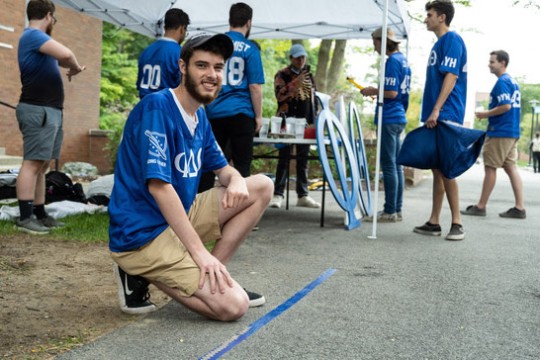 students wearing blue T-shirts standing around a tent as they place quarters on the ground along a piece of tape.