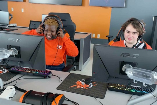 two college students wearing gaming headsets and sitting at a computer station.