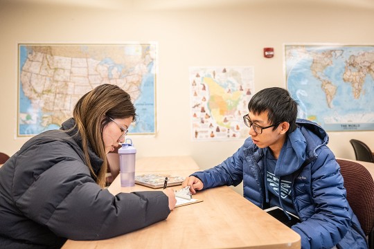 two college students sitting across from each other at a table. There are maps of the United State and the world in the background.